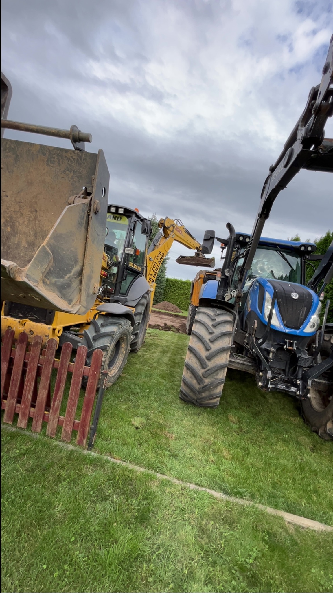 Żółto-czarny traktor z łyżką i niebieski traktor na trawie, obok wykopu. Fragment czerwonego płotu w lewym dolnym rogu. Pochmurne niebo w tle.
