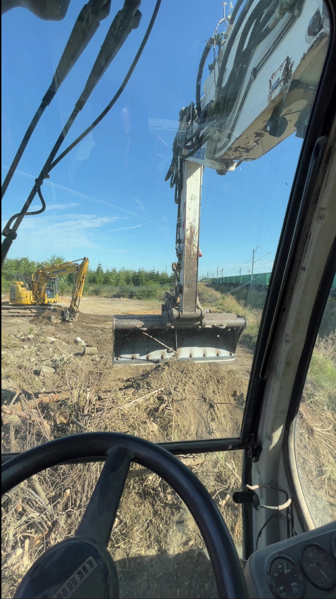 Widok z kabiny na białą koparkę Liebherr podczas prac ziemnych. W tle żółta koparka i linia kolejowa. Prace przygotowawcze pod inwestycję.