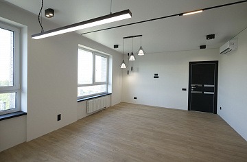 Puste, jasne pomieszczenie po remoncie z parkietem w odcieniach beżu, białymi ścianami, czarnymi akcentami (drzwi, ramy okienne, oświetlenie) i nowoczesnym oświetleniem sufitowym.