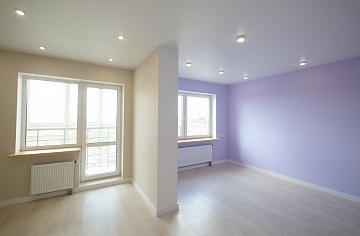 Puste, jasne wnętrze po remoncie: ściana w kolorze liliowym, druga beżowa z oknem, parkiet w odcieniach szarości, widoczne grzejniki i oświetlenie punktowe.