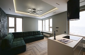 Nowoczesne wnętrze: zielona sofa narożna na parkiecie ułożonym we wzór jodełki, ściana pokryta trójwymiarowymi panelami, minimalistyczna kuchnia z czarnym okapem i oświetleniem LED.