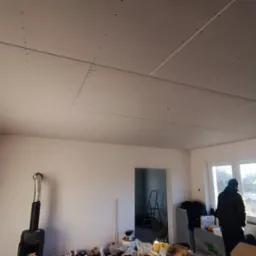 Pokój w trakcie prac wykończeniowych, widoczny zamontowany sufit z płyt karton gips, ślady po szpachlowaniu, osoba w kurtce przy oknie, piec typu koza, bałagan na podłodze.
