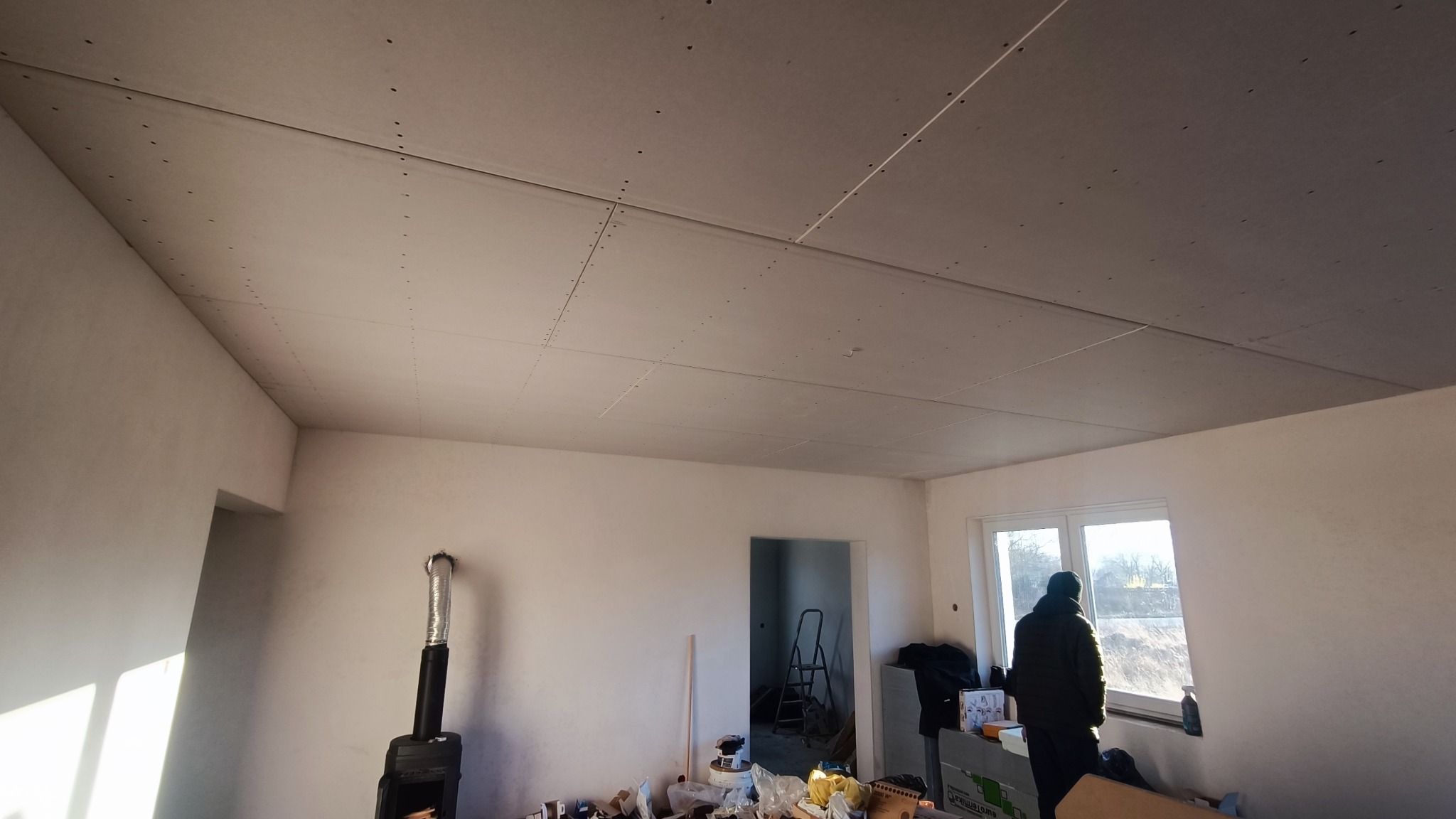 Pokój w trakcie prac wykończeniowych, widoczny zamontowany sufit z płyt karton gips, ślady po szpachlowaniu, osoba w kurtce przy oknie, piec typu koza, bałagan na podłodze.