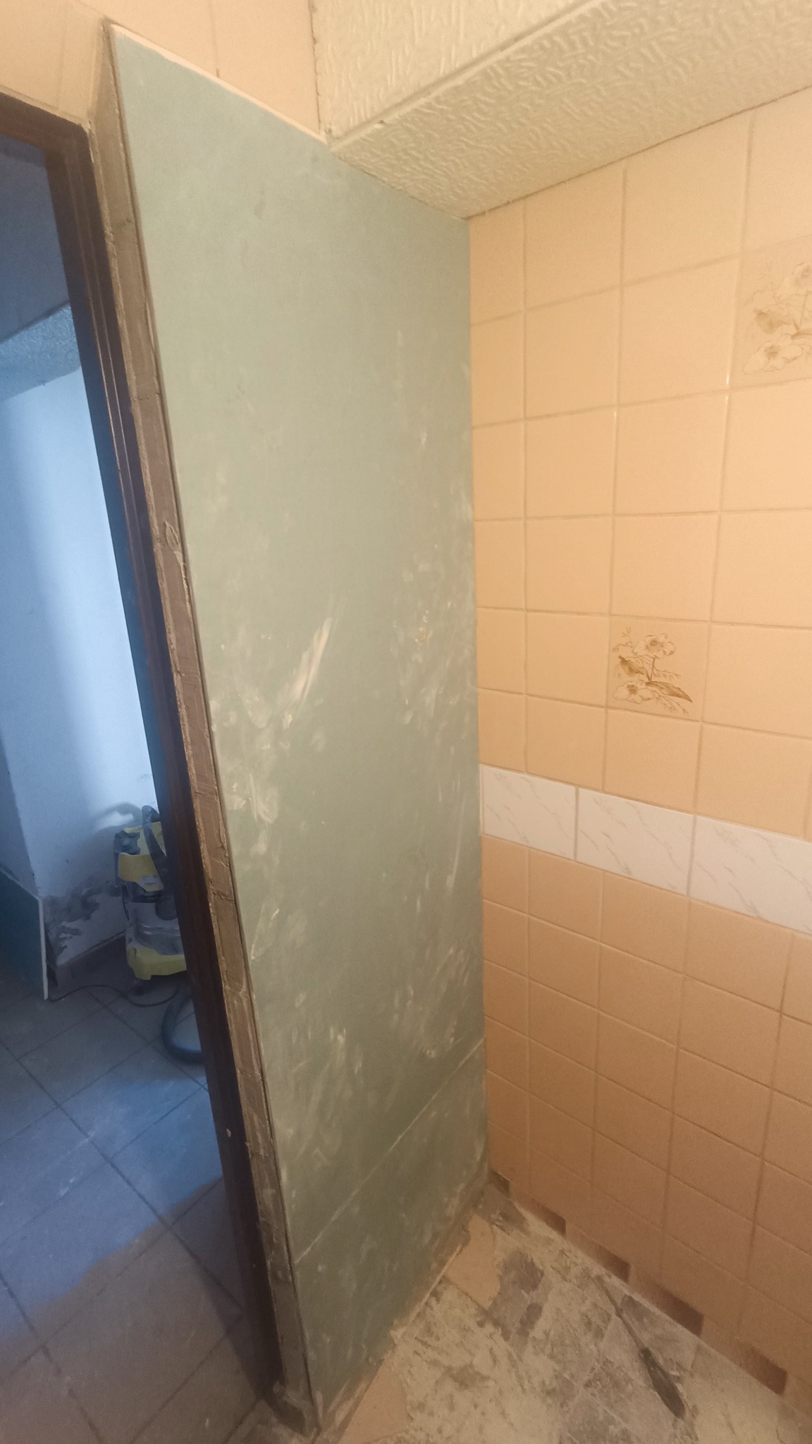 Płyta kartonowo-gipsowa oparta o ścianę z beżowych płytek w trakcie prac wykończeniowych w łazience, widoczny odkurzacz przemysłowy w tle.