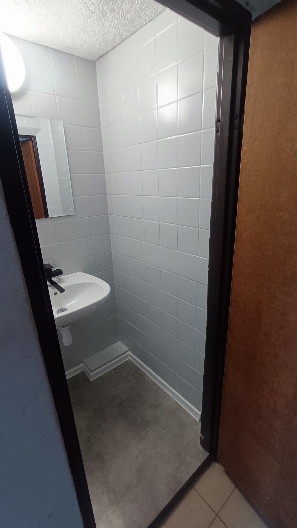 Mała łazienka po remoncie z białymi, kwadratowymi płytkami na ścianach, ciemną podłogą i umywalką z czarną baterią, widok zza framugi drzwi.