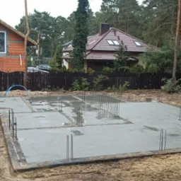 Wylany, świeży fundament pod dom z wystającymi prętami zbrojeniowymi, widok na budynek w trakcie budowy i zalesione otoczenie.