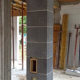 Wznoszenie komina z szarych bloczków betonowych z widocznym otworem na wkład kominkowy, w surowym stanie budynku z widocznymi elementami konstrukcyjnymi.