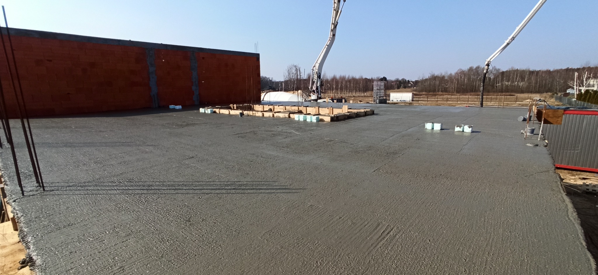 Wylany, świeży beton na dużej powierzchni z wystającymi prętami zbrojeniowymi, częściowo wymurowaną ścianą z czerwonej cegły i betonowymi pompami w tle.