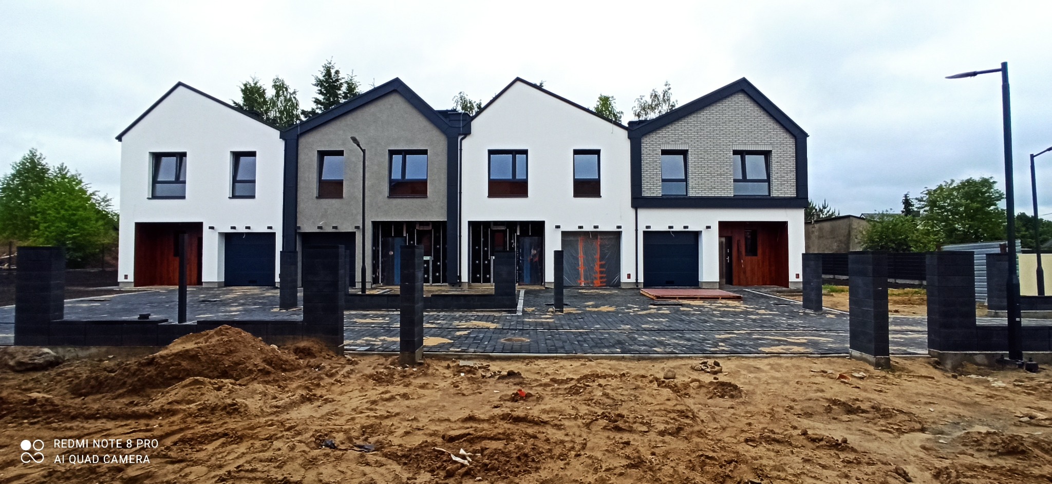 Szeregówka nowych domów jednorodzinnych w trakcie budowy, widoczne fundamenty, mury oraz częściowo ułożona kostka brukowa. Na pierwszym planie hałda ziemi.