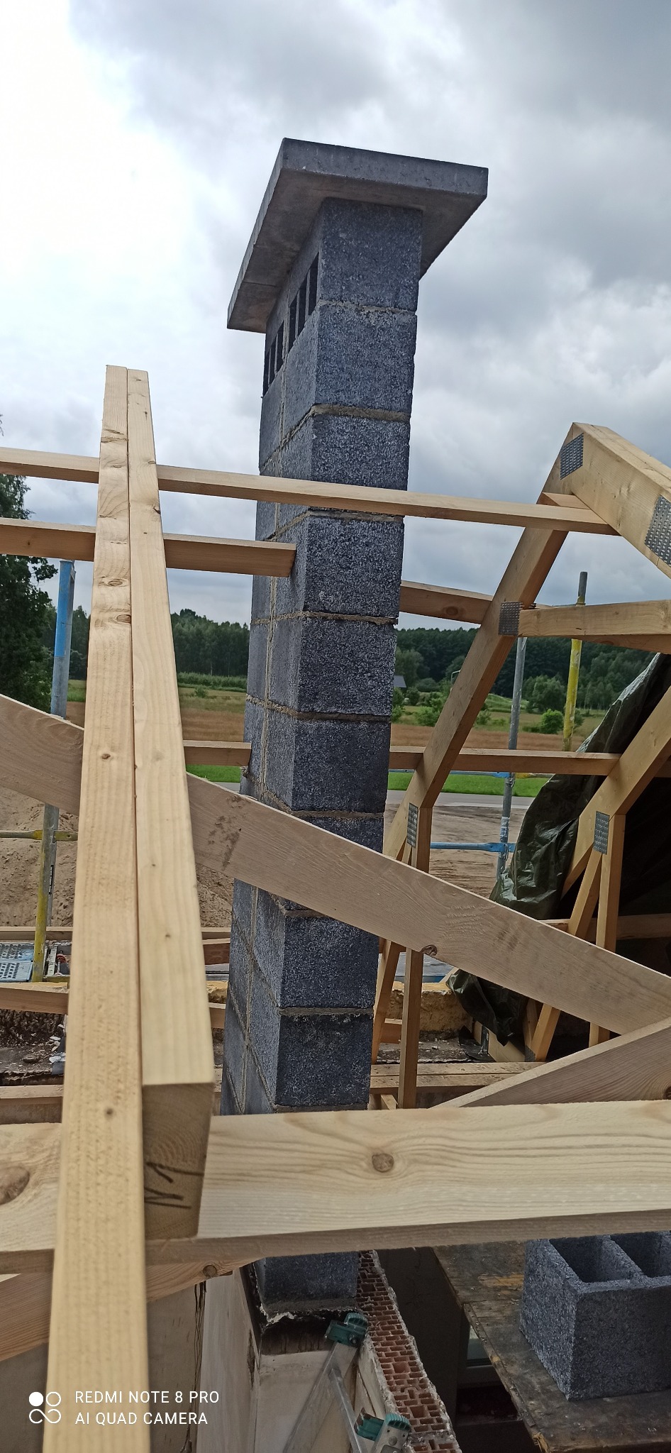 Wznoszenie komina z szarego bloczka betonowego na tle drewnianej konstrukcji dachu w trakcie budowy domu.