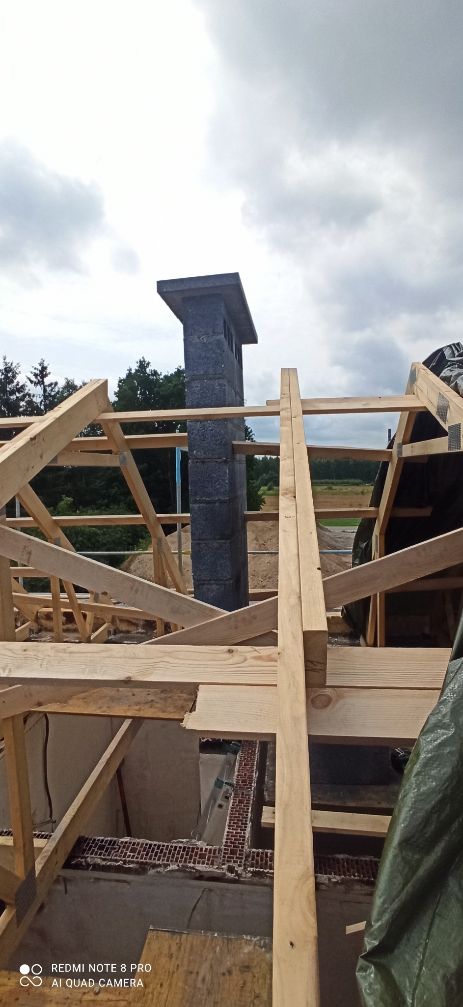 Wznoszenie komina z bloczków na tle drewnianej konstrukcji dachu w trakcie budowy domu, widok z góry na wnętrze budynku.