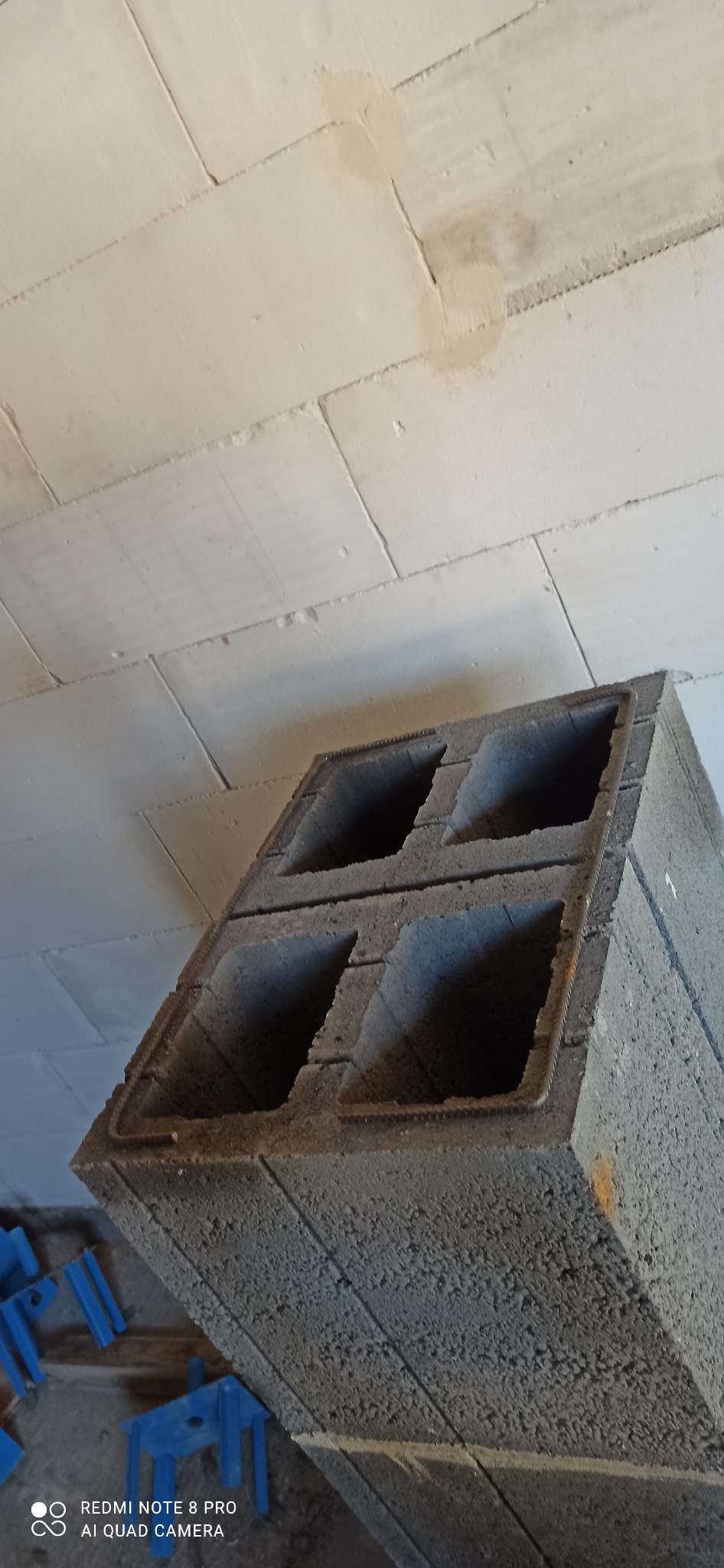 Szary bloczek betonowy z czterema otworami, widoczne zbrojenie, ułożony na ścianie z betonu komórkowego, niebieskie plastikowe elementy stabilizujące w tle.