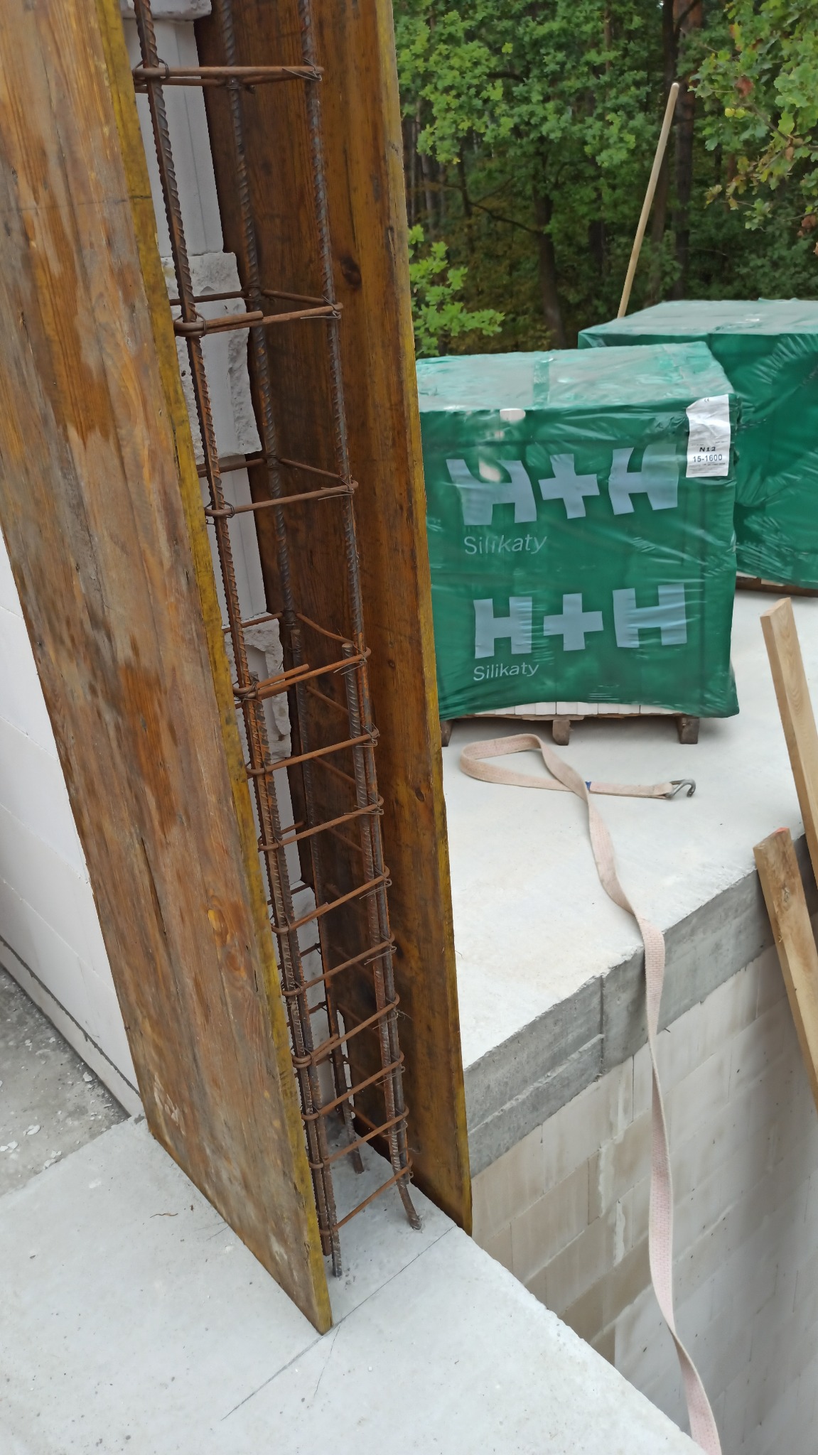 Szczegółowy widok szalunku słupa zbrojonego stalą na fundamencie z bloczków silikatowych, w tle zapakowane bloczki H+H.