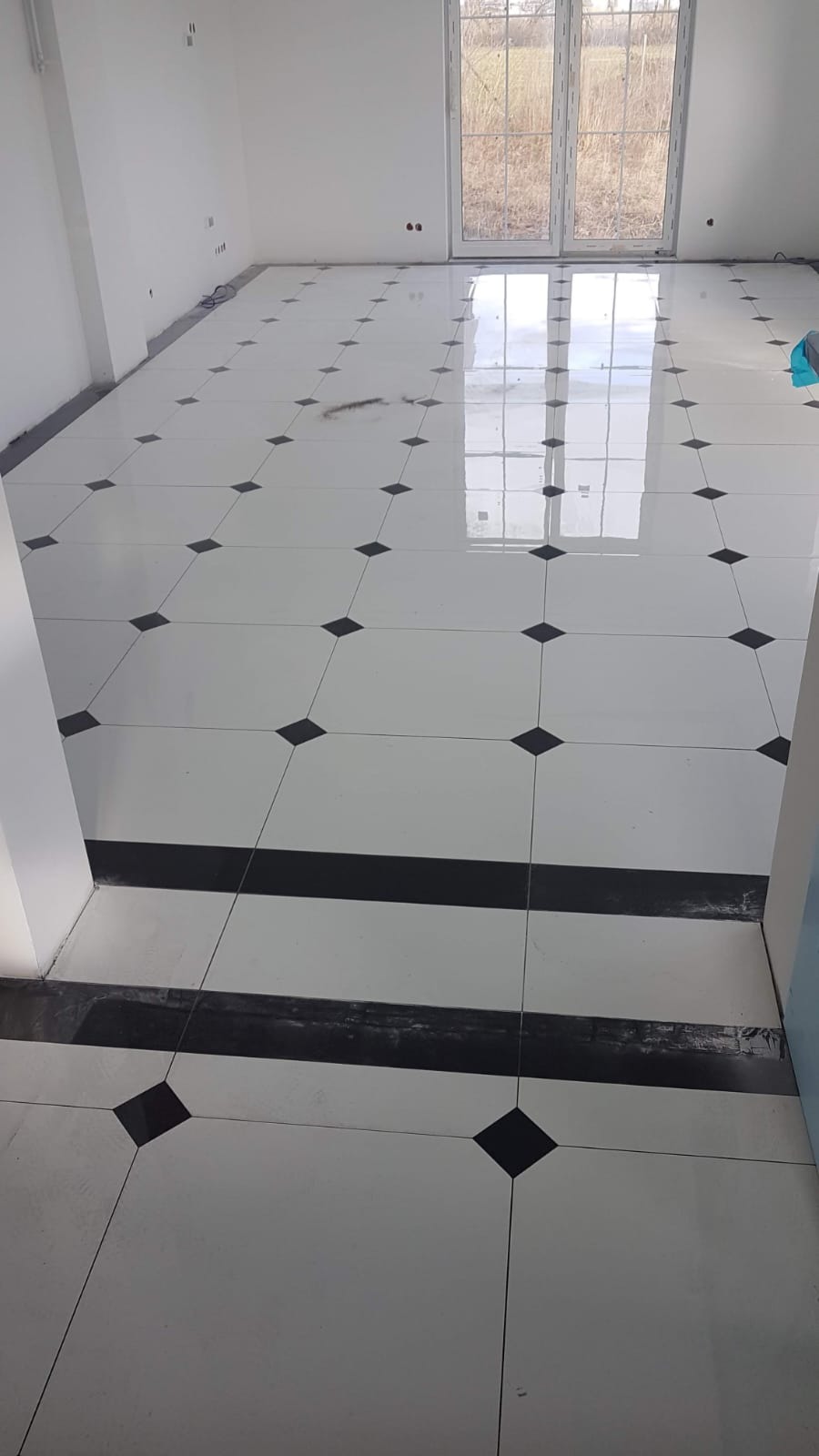 Biała, błyszcząca podłoga wyłożona płytkami z czarnymi rombami w nowym domu, widoczne okno z odbiciem krajobrazu.