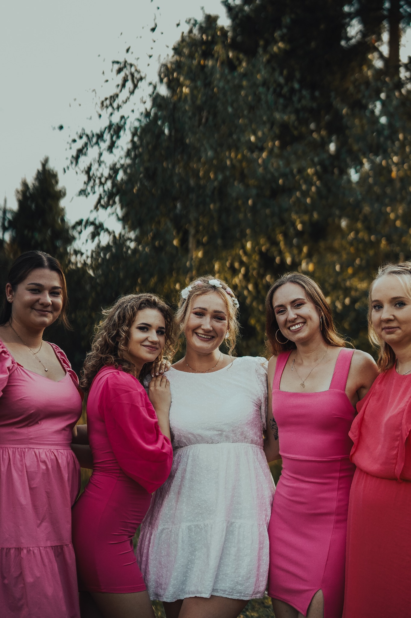 Pięć uśmiechniętych kobiet w różowych sukienkach pozuje na tle zieleni, jedna z nich ma wianek na głowie.