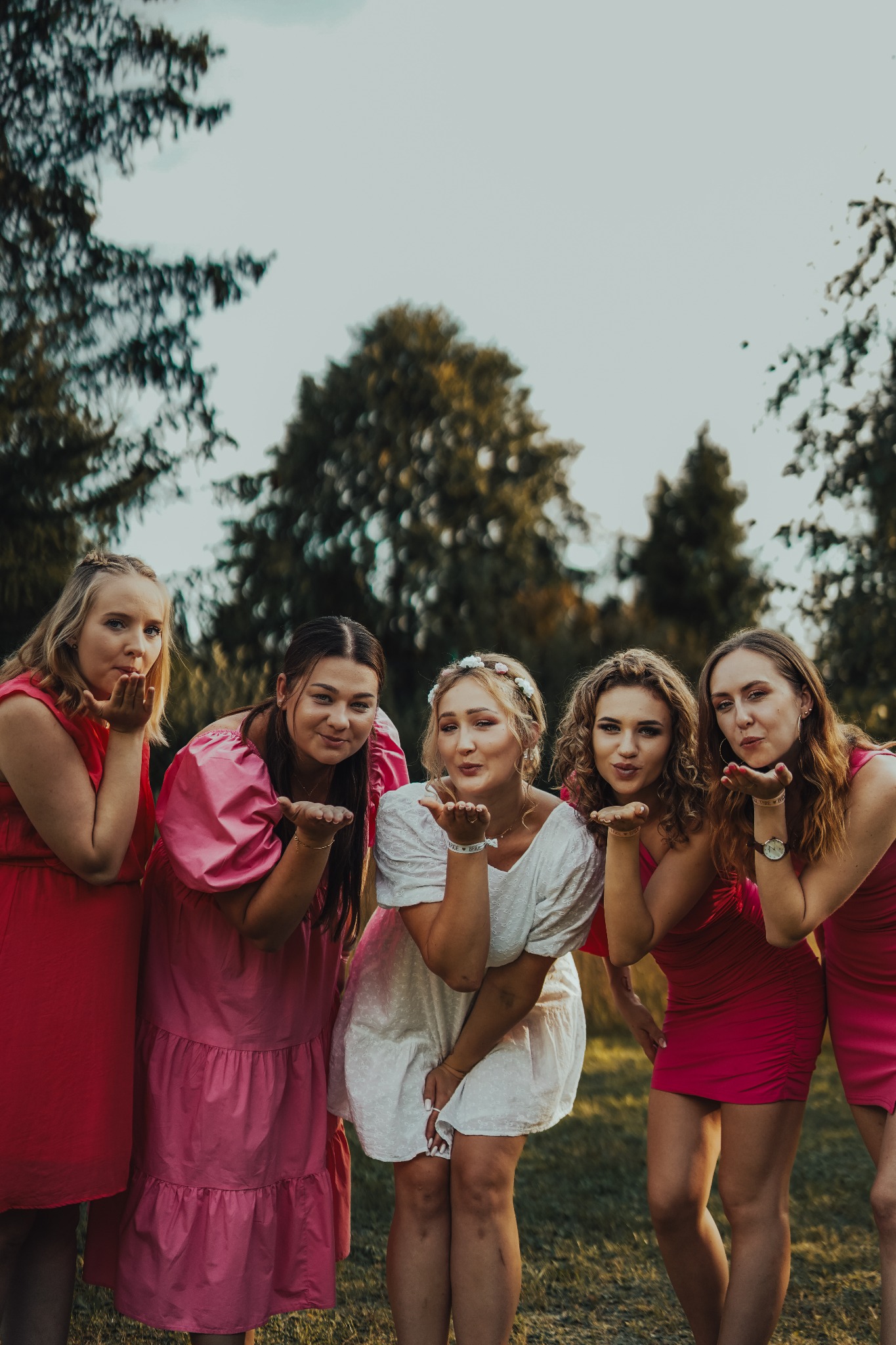 Pięć kobiet w różowych i białych sukienkach dmucha w dłonie na tle zielonych drzew.