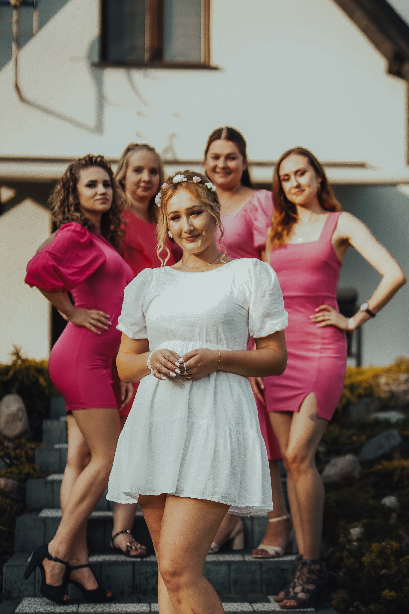 Panna młoda w krótkiej białej sukience i wianku na głowie, pozuje z druhnami w różowych sukienkach na tle białego domu.