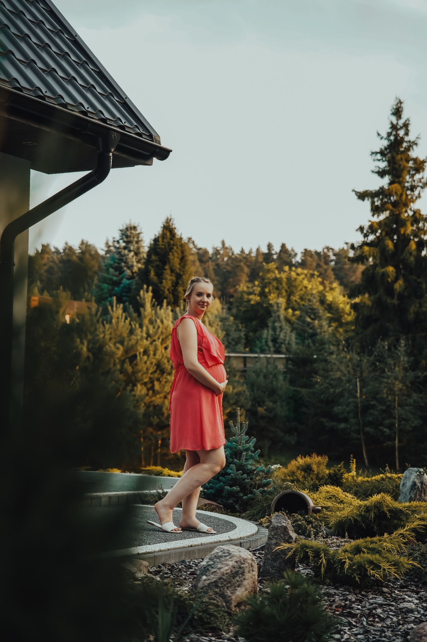 Uśmiechnięta kobieta w koralowej sukience ciążowej, stojąca w ogrodzie na tle domu z ciemnym dachem i zieleni