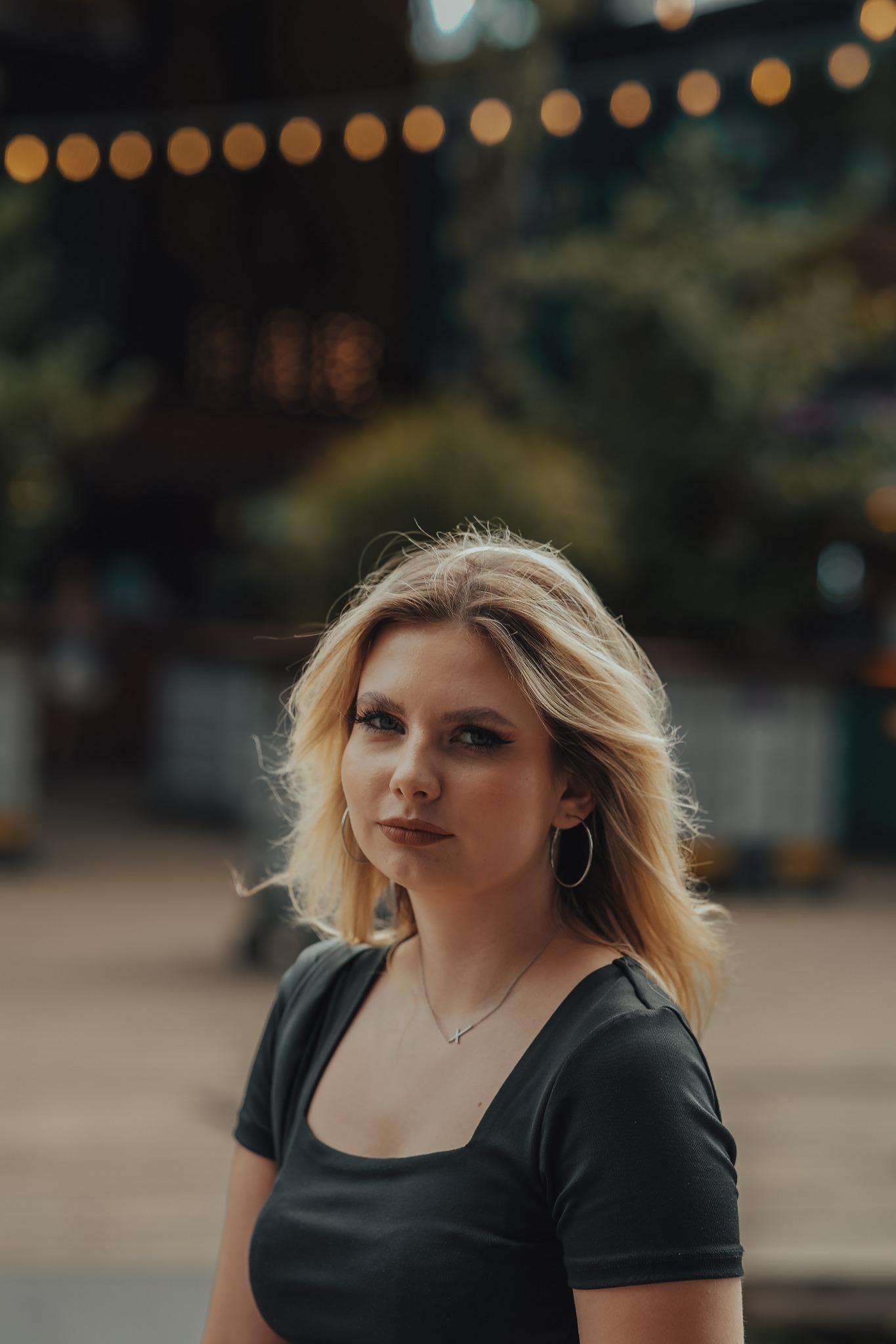 Portret młodej kobiety z blond włosami, srebrnymi kolczykami kołami i naszyjnikiem z krzyżykiem, na rozmytym tle z lampkami.