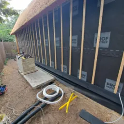 Budowa ściany z trzciny: widoczne drewniane listwy, czarna membrana z nadrukiem 'PROF PARTNER' i izolacja termiczna na fundamencie. Kable i narzędzia leżą na ziemi.