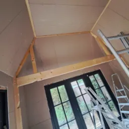 Poddasze z ukośnymi ścianami wykończone płytami kartonowo-gipsowymi, widoczne drewniane belki konstrukcyjne i drabina aluminiowa oparta o ścianę.