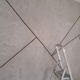 Dekoracyjna ściana z szarym tynkiem strukturalnym i czarnymi, geometrycznymi liniami, częściowo zasłonięta przez aluminiową drabinę.