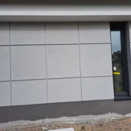 Elewacja budynku z szarego tynku podzielonego na kwadraty, widoczne okno z żółtą naklejką i ciemny cokół, teren wokół budynku w trakcie prac wykończeniowych.