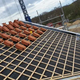 Układanie dachówki ceramicznej na konstrukcji drewnianej z membraną, widoczny żuraw budowlany i rusztowanie w tle.