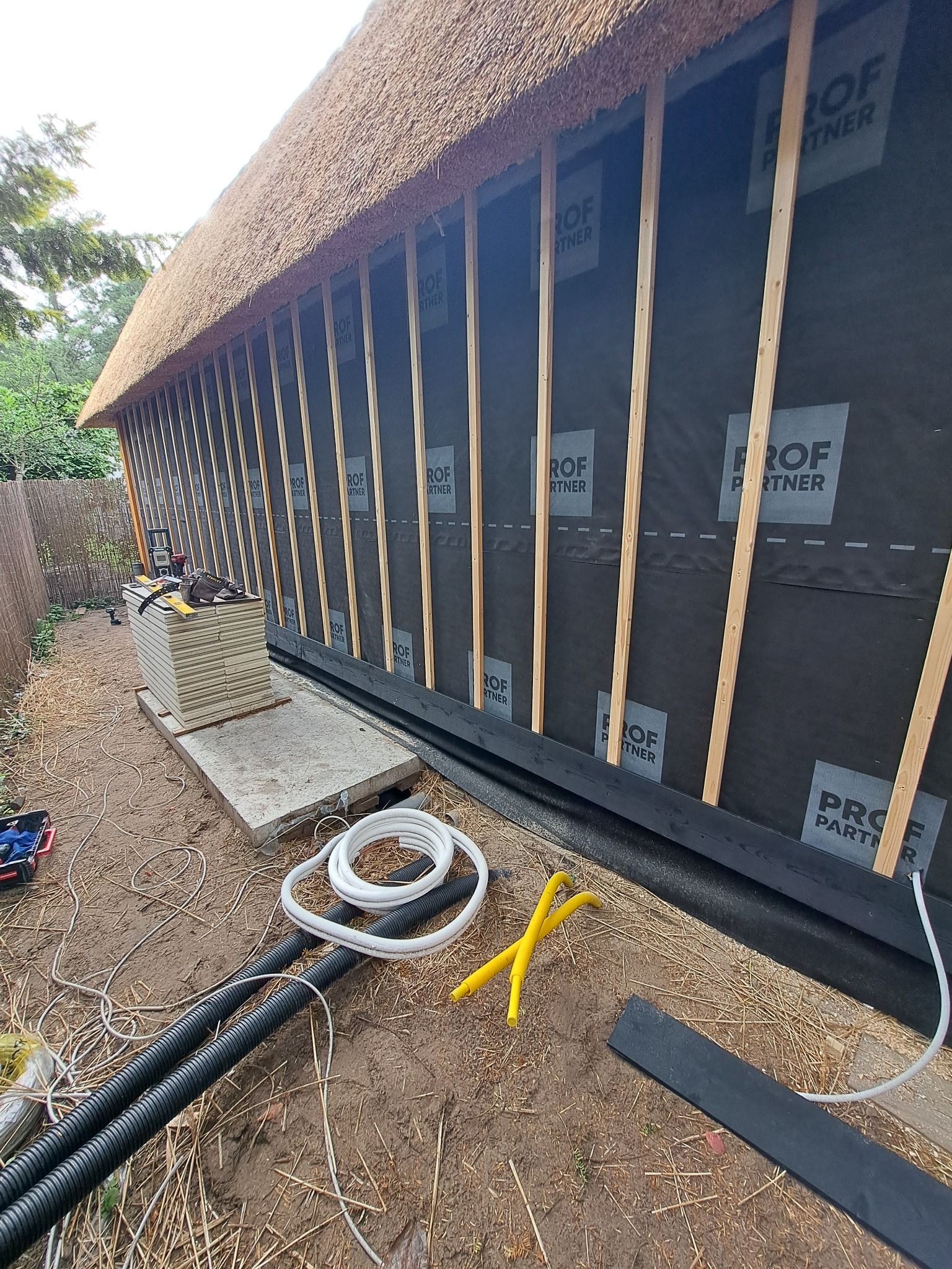 Budowa ściany z trzciny: widoczne drewniane listwy, czarna membrana z nadrukiem 'PROF PARTNER' i izolacja termiczna na fundamencie. Kable i narzędzia leżą na ziemi.