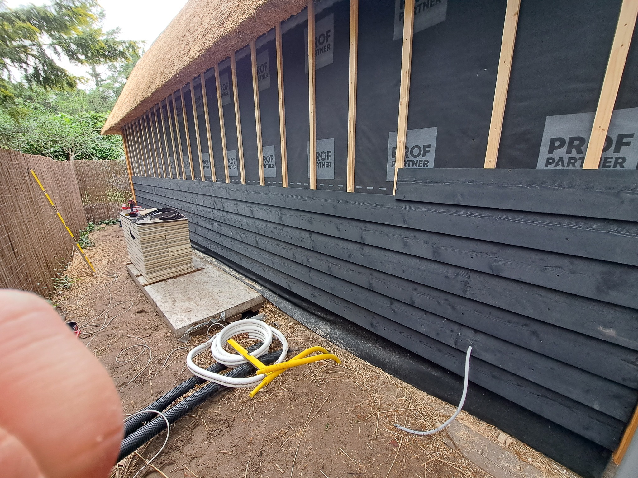 Budynek z czarną drewnianą elewacją i strzechą w trakcie prac: widoczne ocieplenie, drewniane listwy montażowe, membrany z nadrukiem, narzędzia i kable na ziemi.