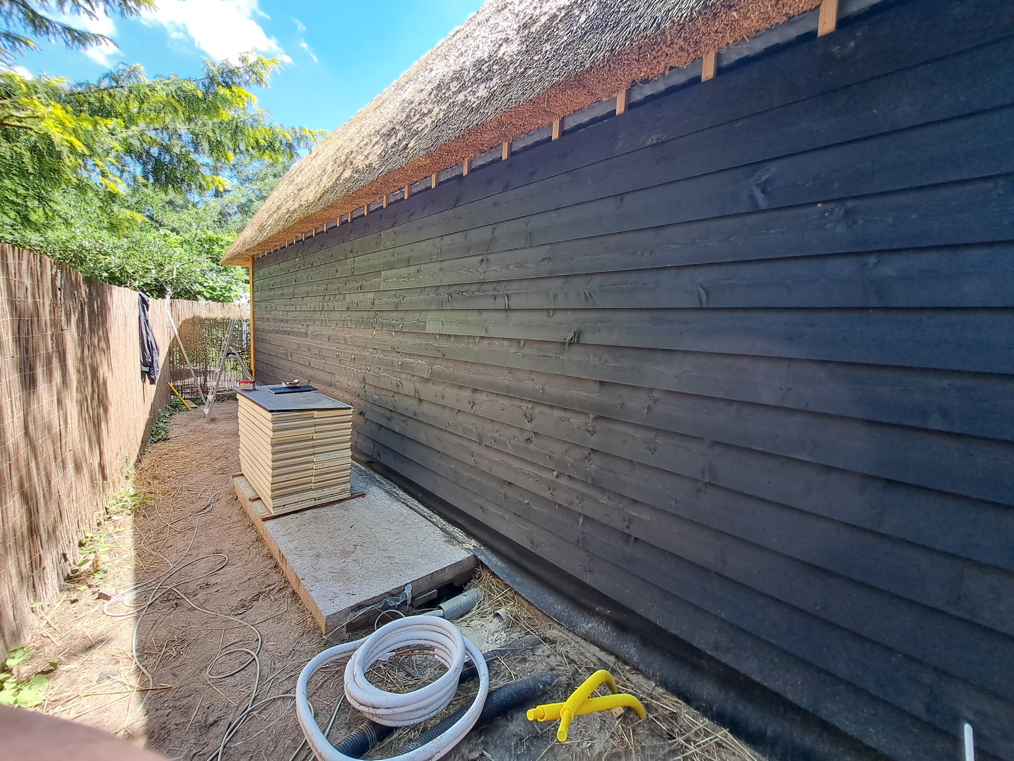 Czarna drewniana elewacja budynku z strzechą, widoczne przygotowane płyty izolacyjne i przewody elektryczne na ziemi.