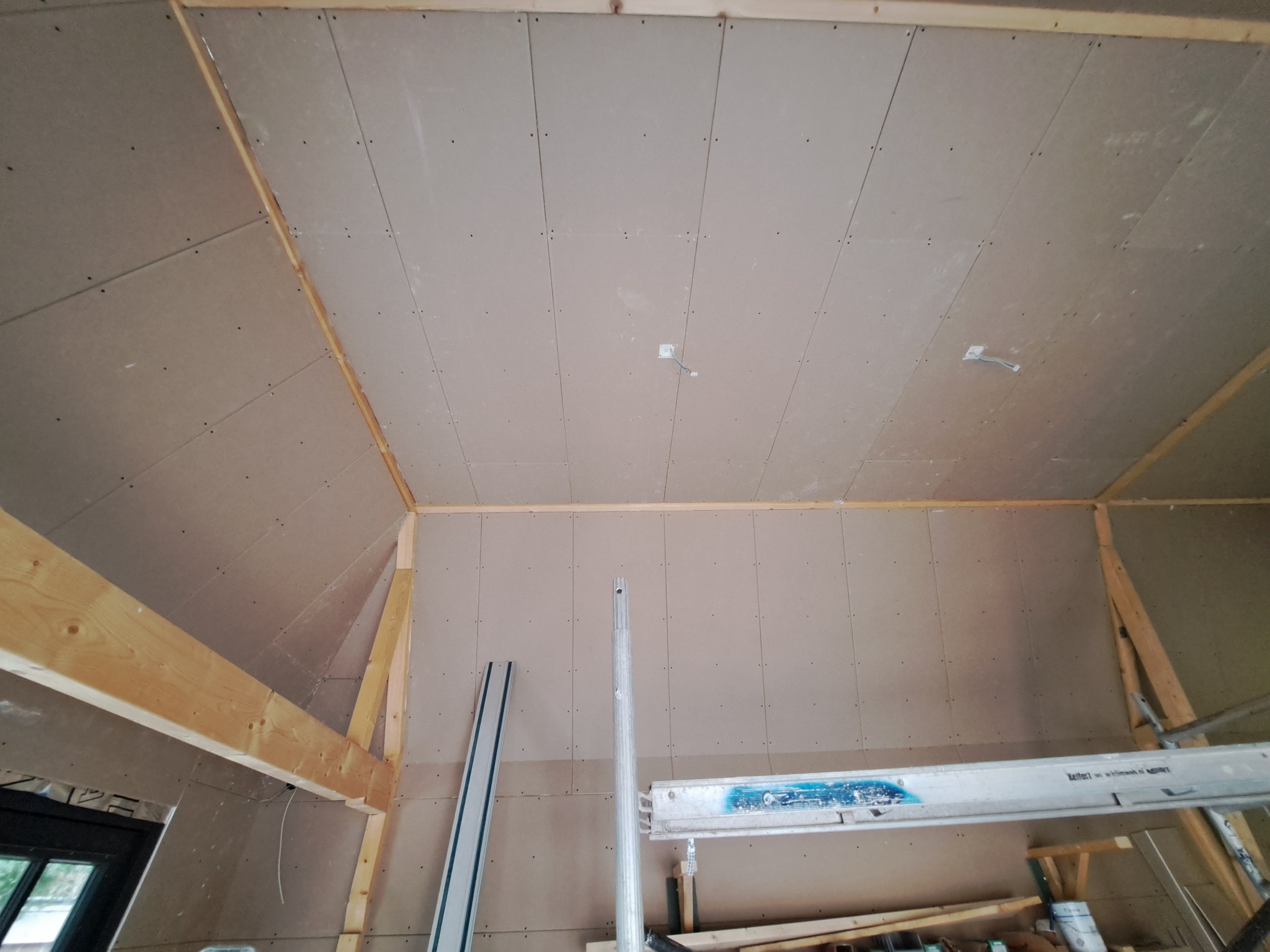 Poddasze w trakcie wykańczania: zamontowane płyty kartonowo-gipsowe na ścianach skośnych i suficie, widoczne drewniane elementy konstrukcyjne, fragment okna, drabina i profile aluminiowe.
