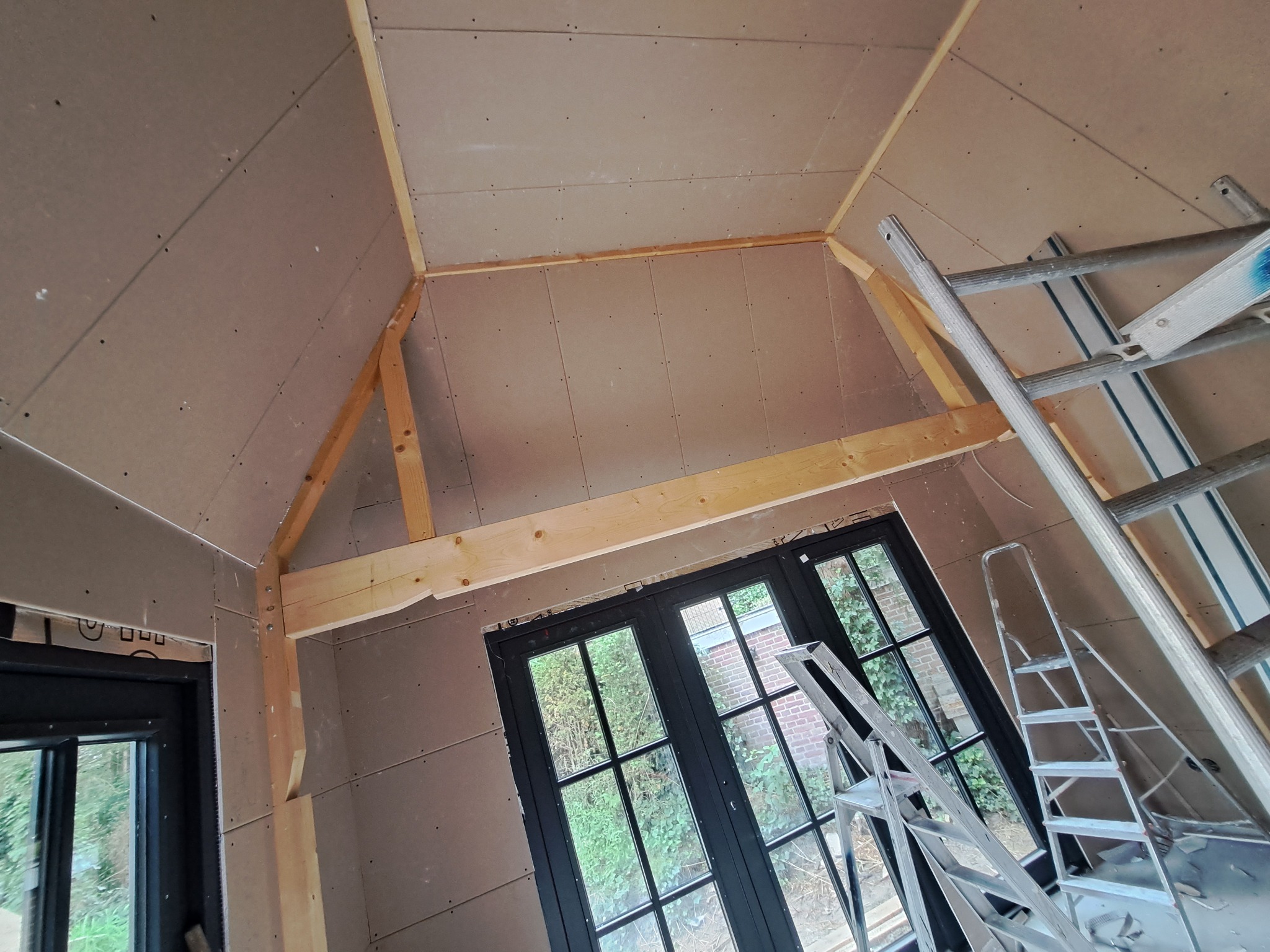 Poddasze z ukośnymi ścianami wykończone płytami kartonowo-gipsowymi, widoczne drewniane belki konstrukcyjne i drabina aluminiowa oparta o ścianę.