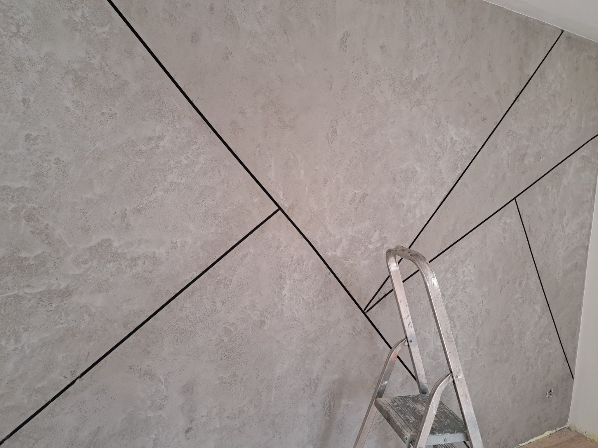 Dekoracyjna ściana z szarym tynkiem strukturalnym i czarnymi, geometrycznymi liniami, częściowo zasłonięta przez aluminiową drabinę.