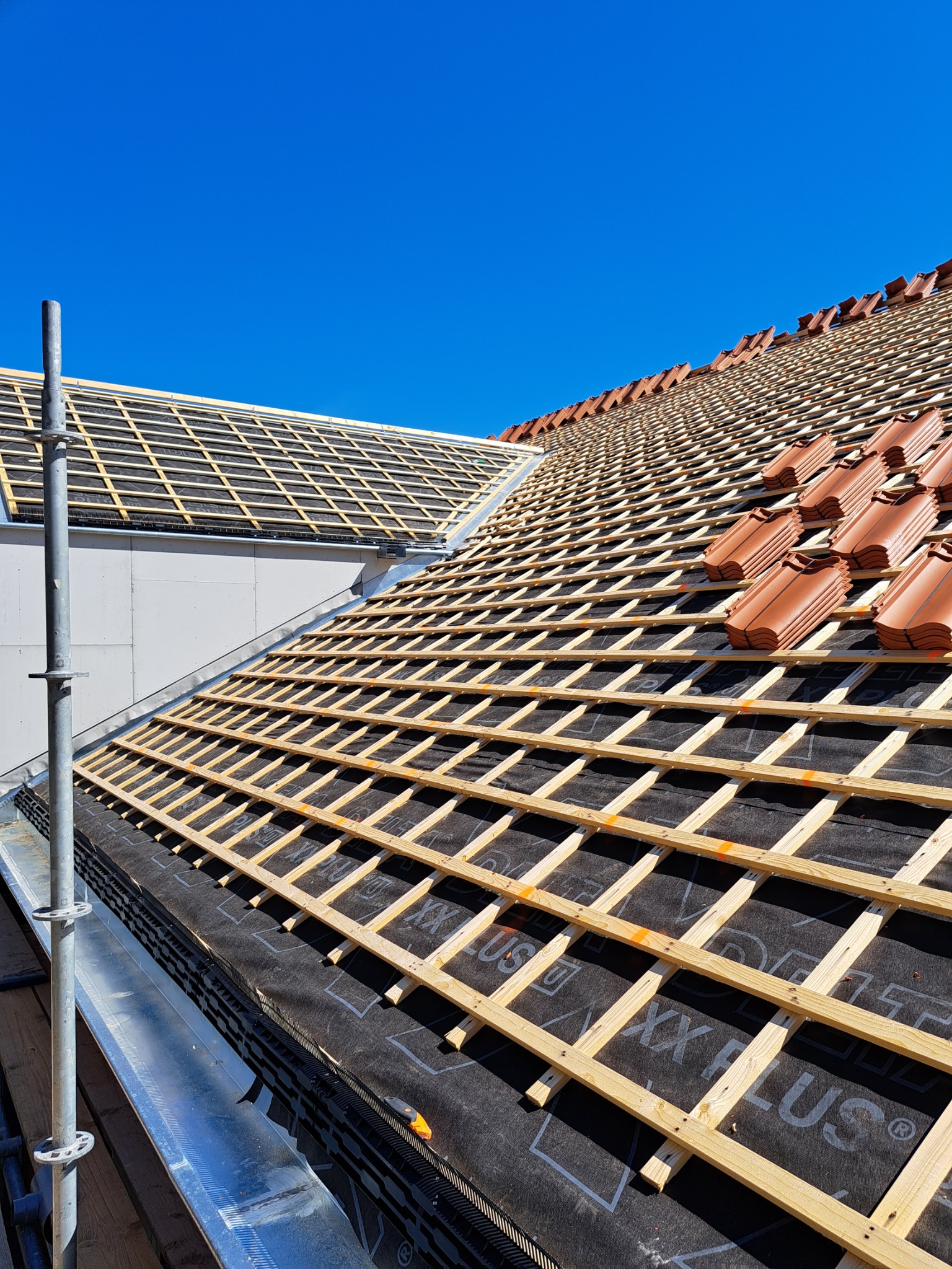 Montaż łat i kontrłat na dachu pokrytym membraną, częściowo ułożona dachówka ceramiczna, widoczne rusztowanie i błękitne niebo.