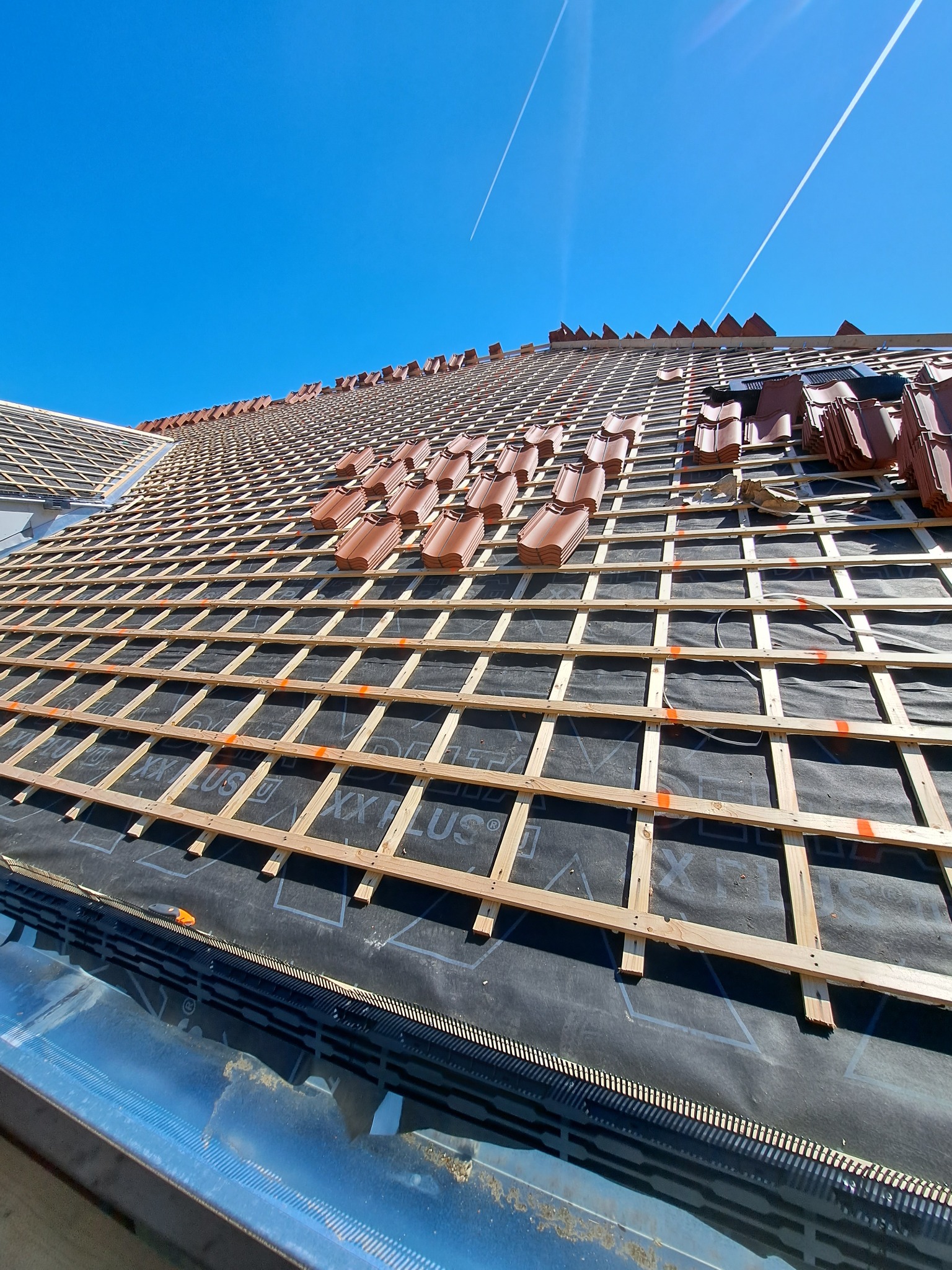 Montaż brązowej dachówki na drewnianej konstrukcji dachu, widoczne membrany dachowe i błękitne niebo w tle.