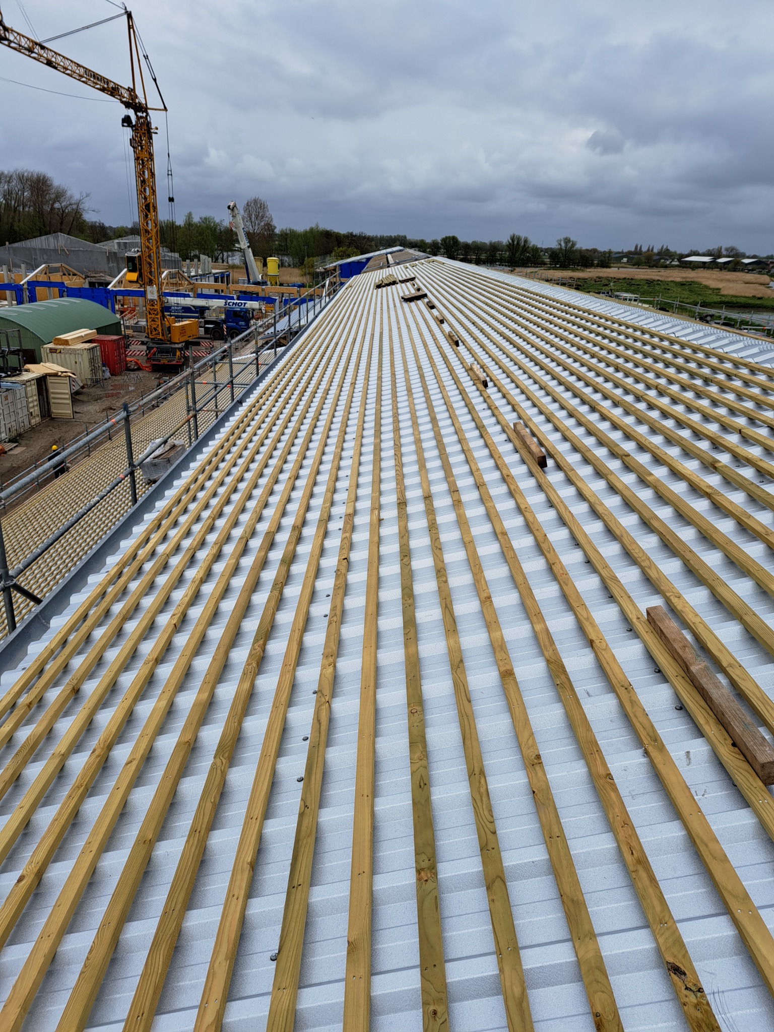 Montaż drewnianej konstrukcji dachu na blaszanym pokryciu, widok z góry, w tle żuraw budowlany i pochmurne niebo.