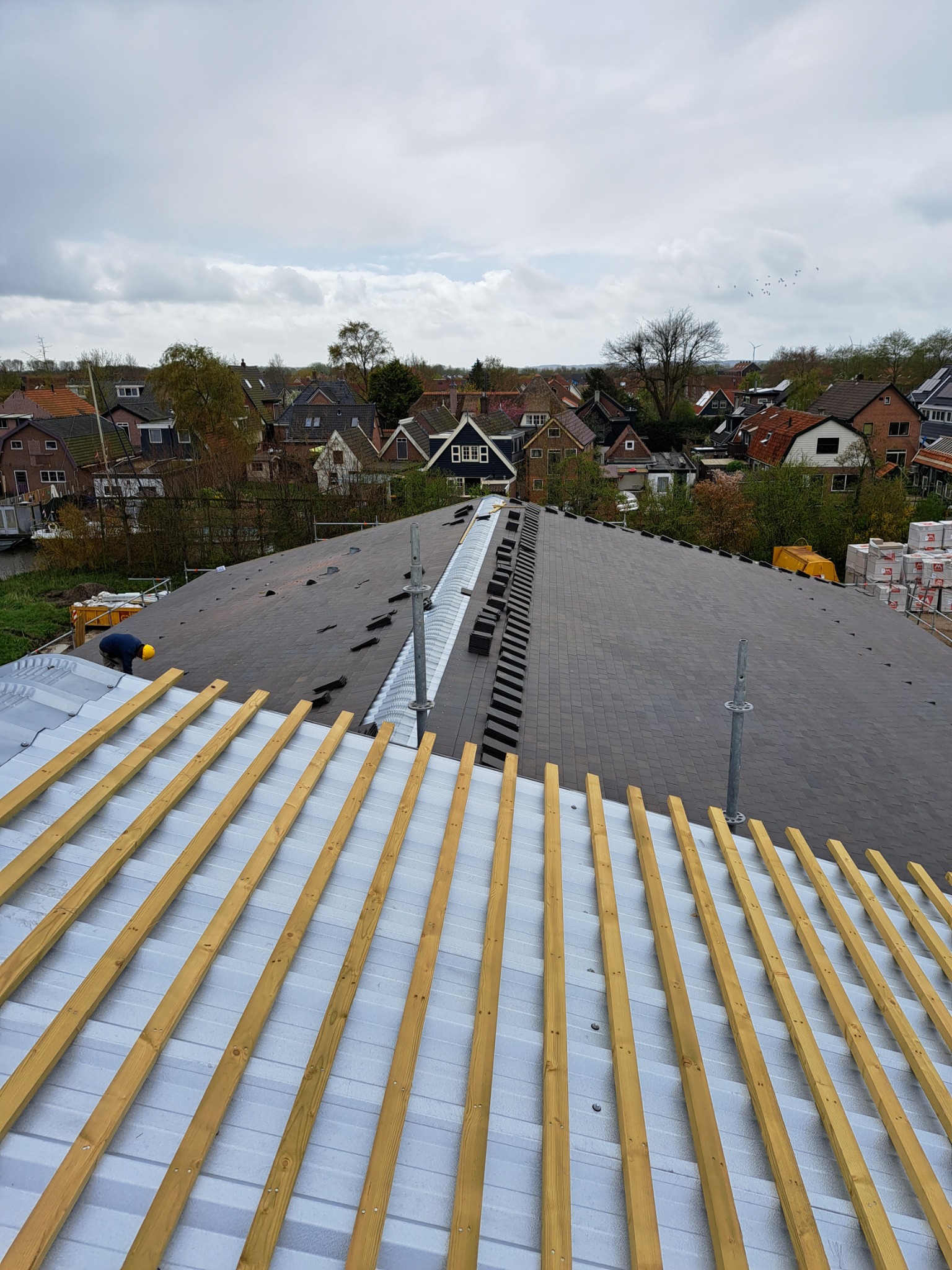 Montaż pokrycia dachowego z dachówki bitumicznej na nowym dachu, widok z góry na częściowo ukończoną powierzchnię z widocznymi łatami i pracownikiem w żółtym kasku.