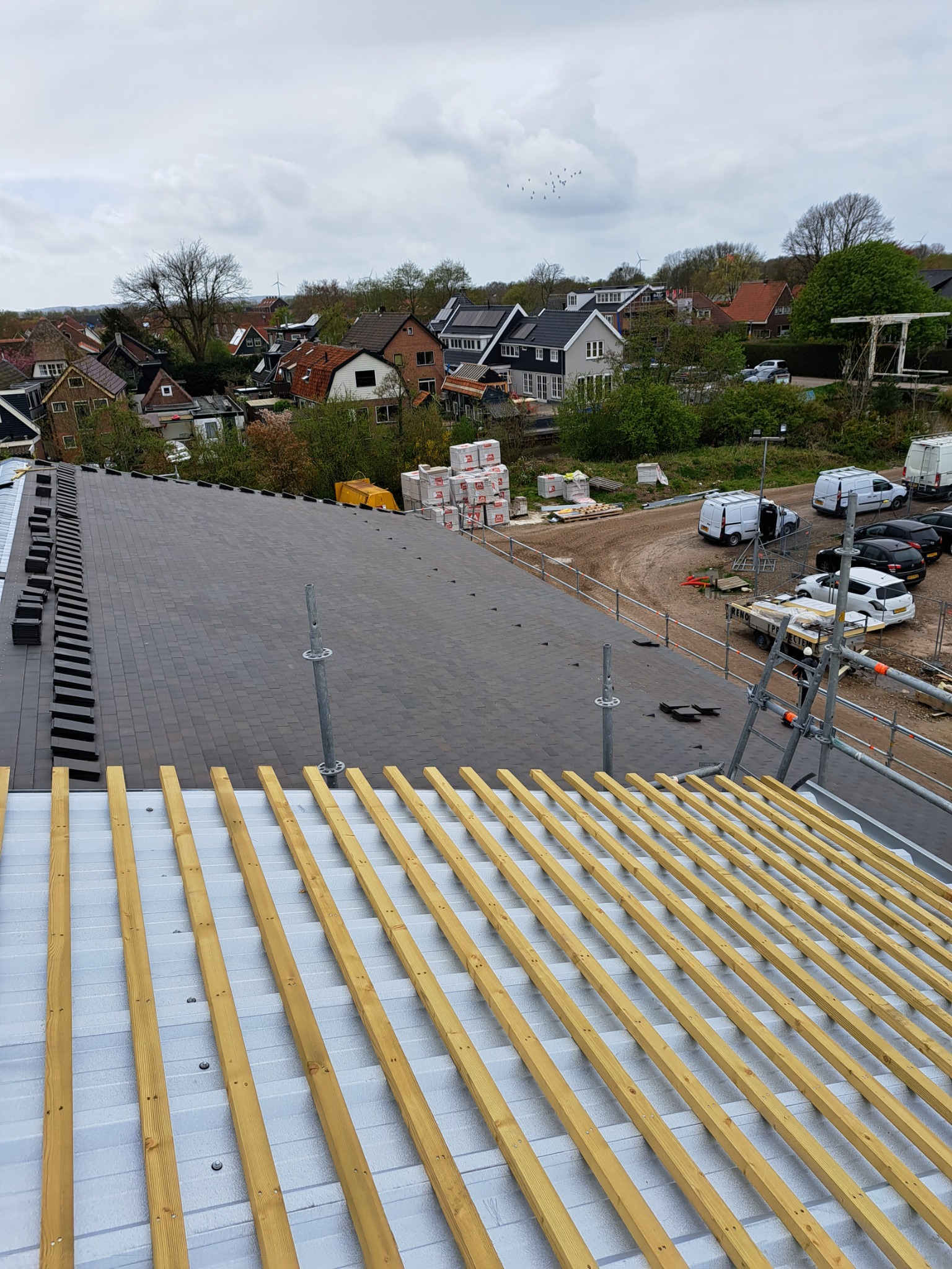 Montaż łat pod dachówkę na metalowej konstrukcji, w tle widok na zabudowę jednorodzinną i pochmurne niebo.