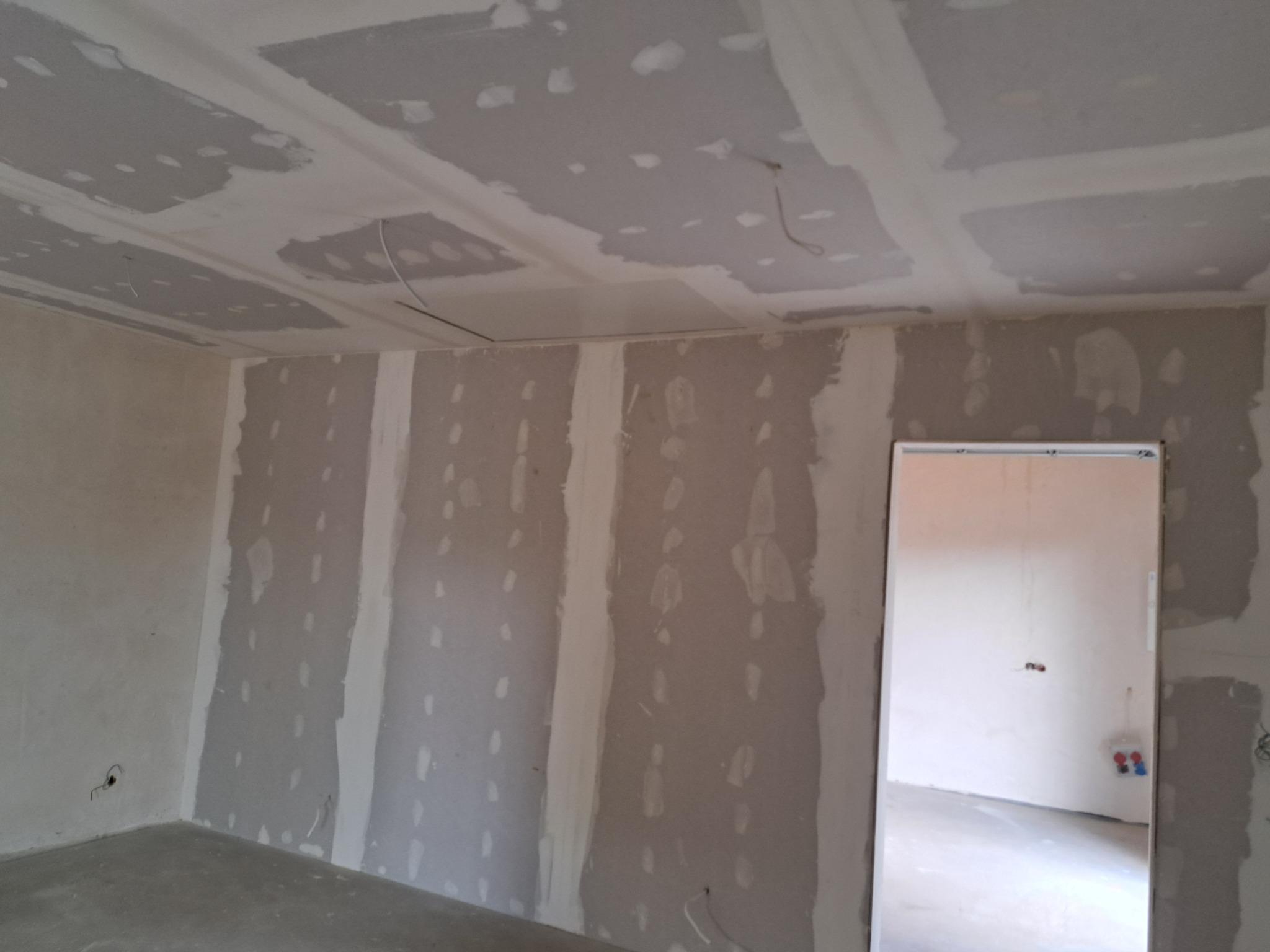 Wnętrze pokoju w trakcie montażu płyt gipsowo-kartonowych na ścianach i suficie, widoczne łączenia płyt pokryte masą szpachlową, surowy betonowy podkład podłogowy, w tle ościeżnica drzwiowa.