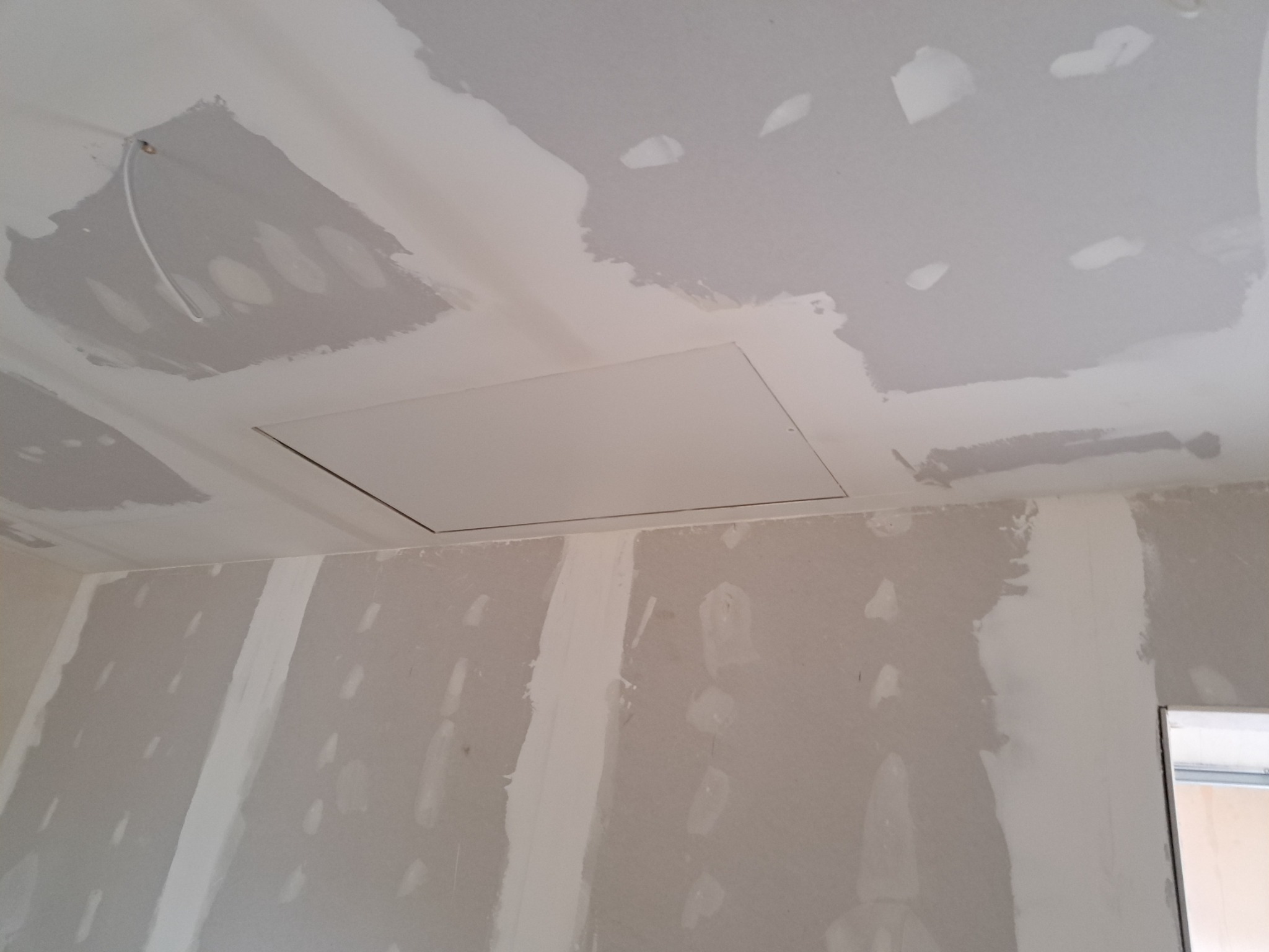 Wnętrze w trakcie prac wykończeniowych: ściany i sufit pokryte płytami gipsowo-kartonowymi, widoczne łączenia szpachlowane, klapa rewizyjna w suficie, instalacja elektryczna wystająca z sufitu.