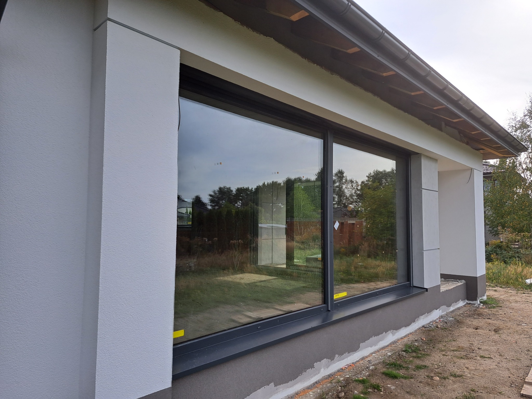 Fragment elewacji domu jednorodzinnego z dużym oknem przesuwnym, widoczne wykończenie tynkiem strukturalnym w jasnym odcieniu szarości oraz ciemnoszara podmurówka w trakcie wykańczania.