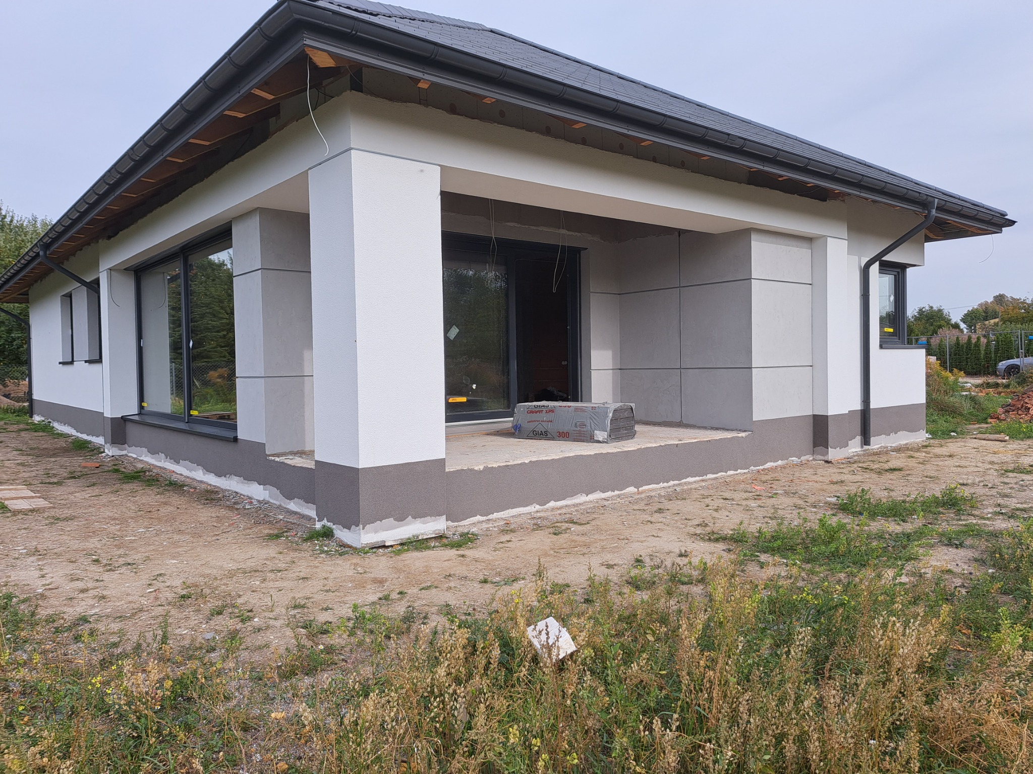 Niedokończony parterowy dom z szarą elewacją, widocznymi oknami, czarną rynną i kostką termoizolacyjną na tarasie.