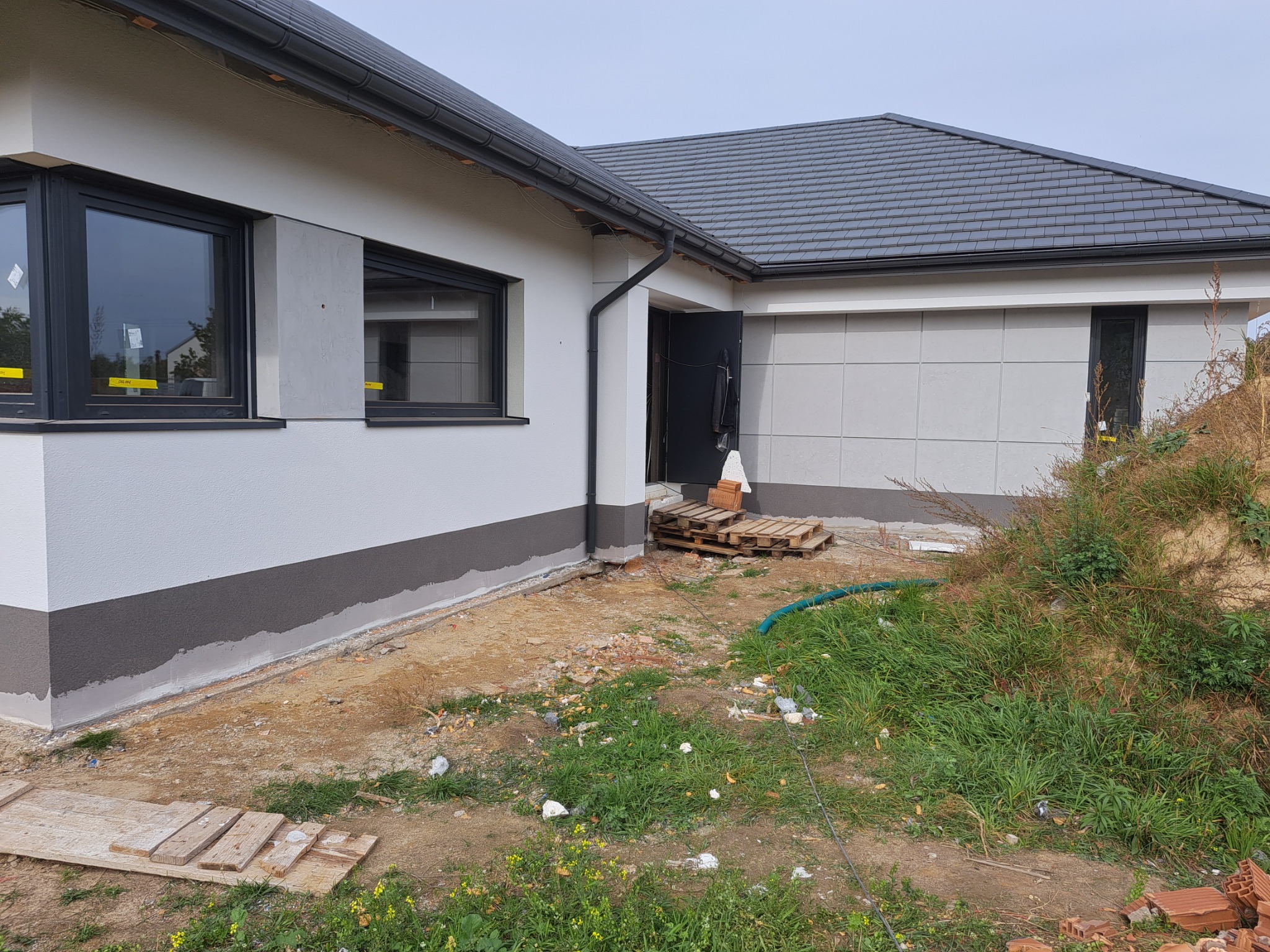 Nowoczesny dom jednorodzinny w Tomaszowie Mazowieckim z wykończoną elewacją w odcieniach bieli i szarości, widoczne detale wykończeniowe i częściowo zagospodarowany teren wokół budynku.