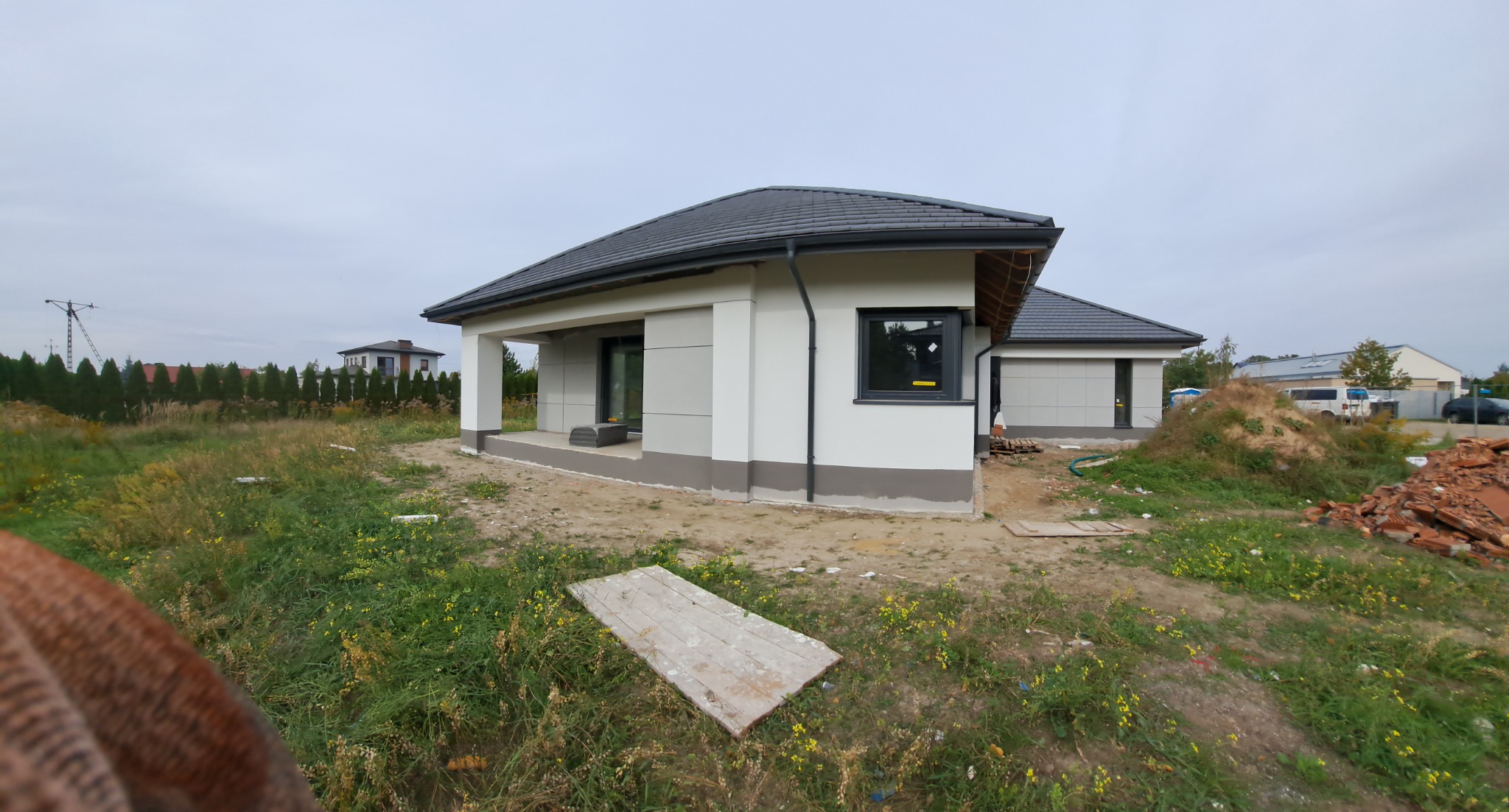 Nowoczesny dom jednorodzinny z płaskim dachem, otynkowany na biało z szarym cokołem, widok z zewnątrz na niezagospodarowaną działkę z pozostałościami budowlanymi.