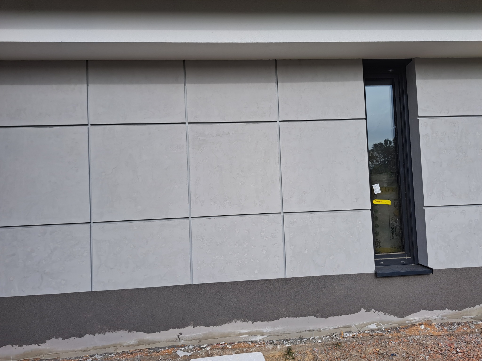 Elewacja budynku z szarego tynku podzielonego na kwadraty, widoczne okno z żółtą naklejką i ciemny cokół, teren wokół budynku w trakcie prac wykończeniowych.
