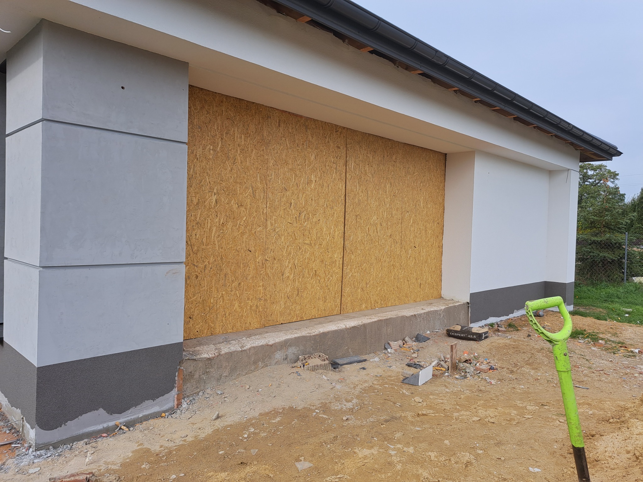 Budynek w trakcie prac elewacyjnych, okna zabezpieczone płytami OSB, widoczny fragment ocieplonego cokołu i zielona łopata.