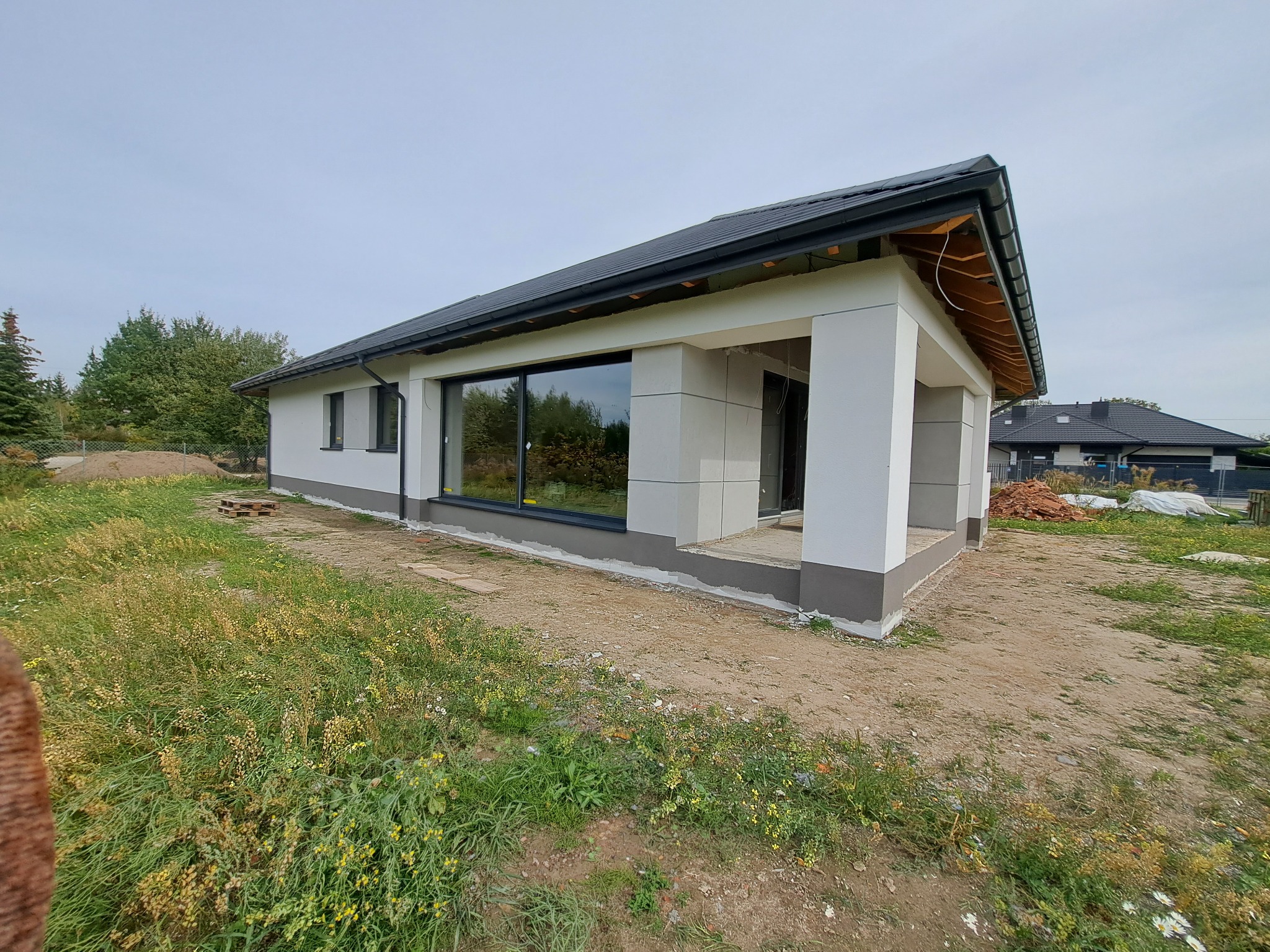 Niedokończony, parterowy dom jednorodzinny z białymi ścianami i szarą podmurówką, widoczne elementy konstrukcyjne werandy, otoczenie w stanie surowym.