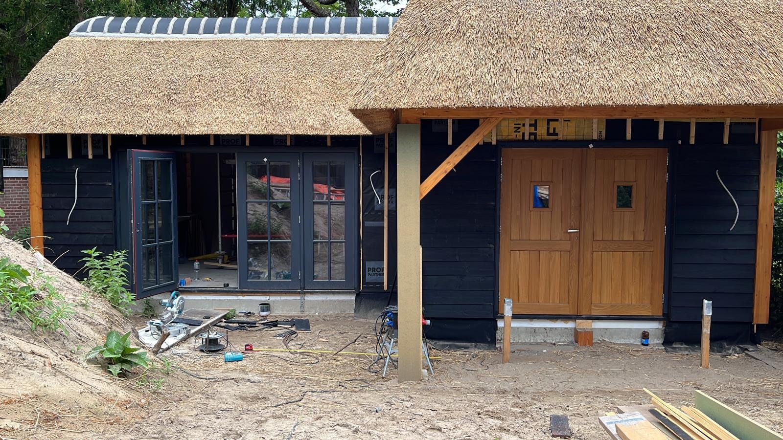 Budynek z czarną drewnianą elewacją i strzechą w trakcie budowy, widoczne narzędzia i materiały budowlane na zewnątrz.