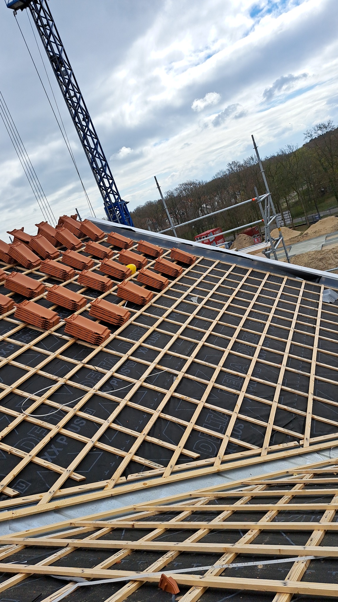 Układanie dachówki ceramicznej na konstrukcji drewnianej z membraną, widoczny żuraw budowlany i rusztowanie w tle.